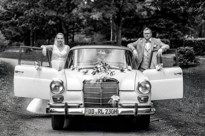 Brautpaar steht neben einem klassischem, weißen Auto auf einem Waldweg.