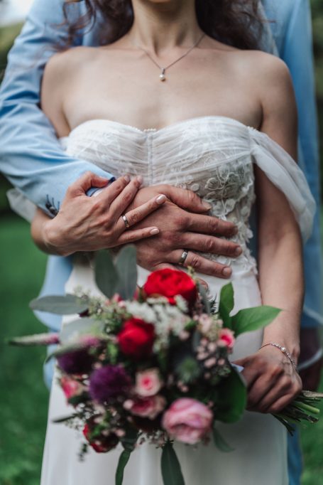 Braut in einem Hochzeitskleid hält einen Blumenstrauß, umarmt von einem Partner.