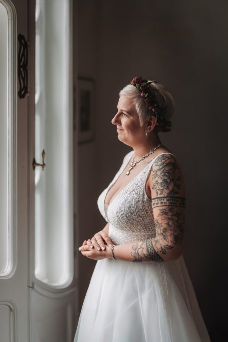 Hochzeitsbraut in einem eleganten Kleid, mit tätowierten Armen, blickt nachdenklich aus dem Fenster.