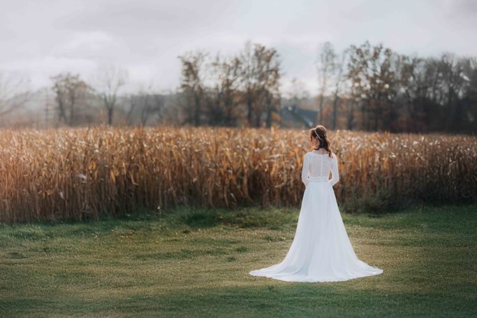 Braut in einem weißen Kleid steht auf einer Wiese vor einem Feld und Bäumen im Hintergrund.