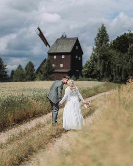 Ein Paar in Hochzeitskleidung geht auf einem Weg zu einer Mühle in einer ländlichen Umgebung.