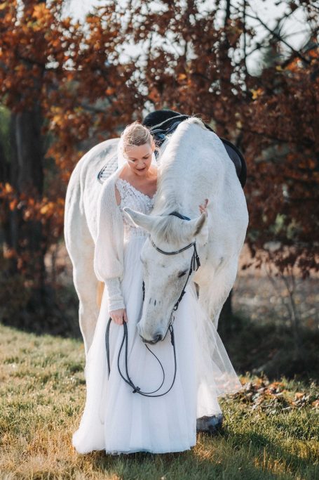 Eine Frau in einem weißen Kleid steht neben einem weißen Pferd in einer herbstlichen Umgebung.