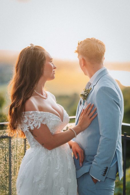 Hochzeitspar auf einem Balkon, Sonnenuntergang im Hintergrund. Brautkleid und Anzug sichtbar.